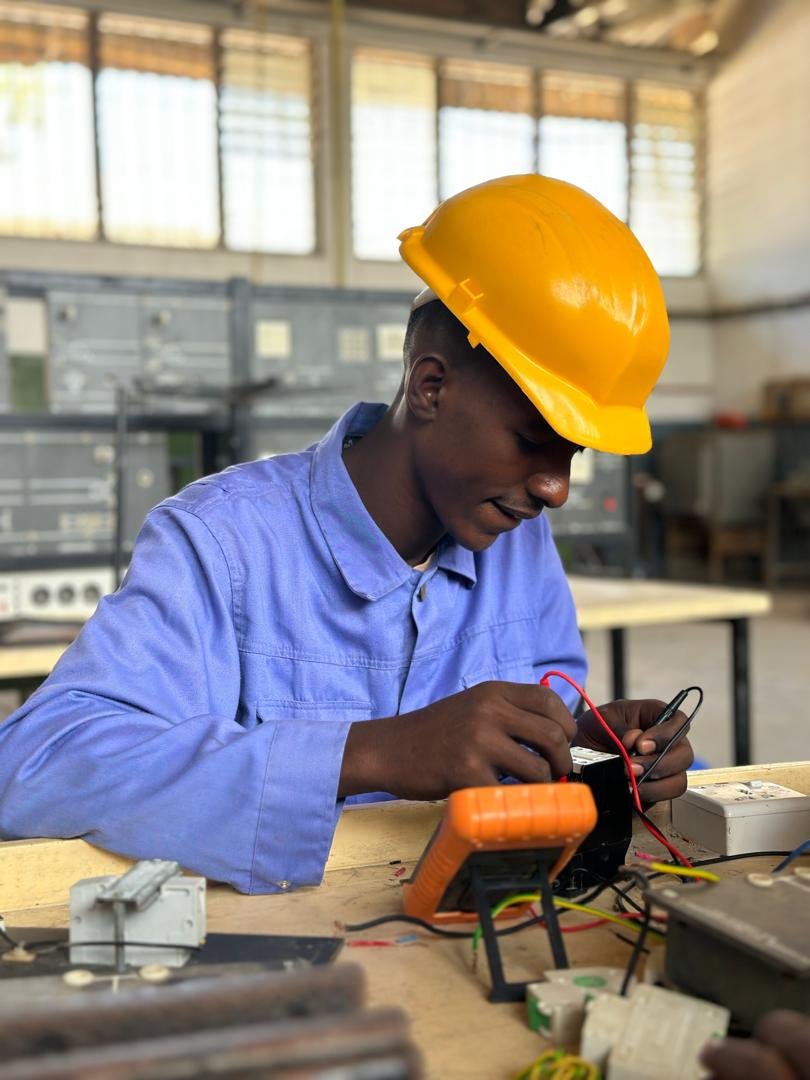 Two students in yellow hard hats and blue overalls working together on an electrical training panel in a workshop