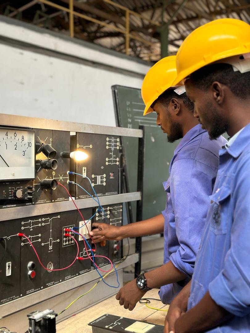 TVET trainee in a yellow hard hat and blue work shirt using a multimeter on electrical equipment